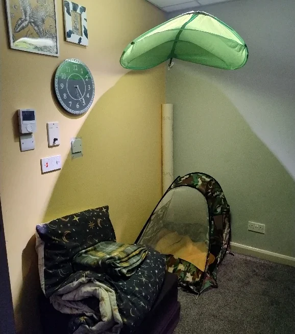 A cozy room corner with a chair, wall clock, hangable umbrella light, and a small tent.