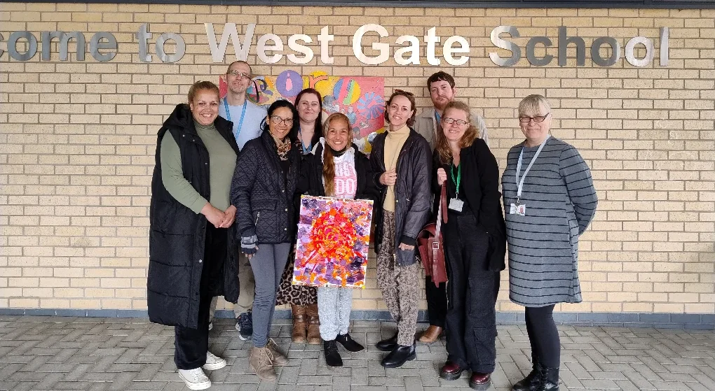 A diverse group of individuals gathered in front of a West Gate School sign