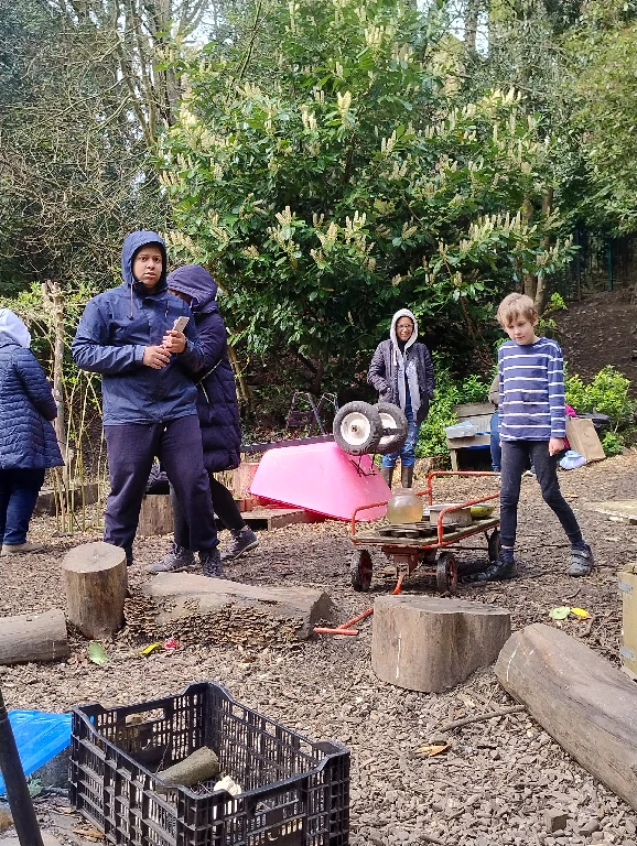 People in an outdoor play area with wooden stumps and a toy wheelbarrow.