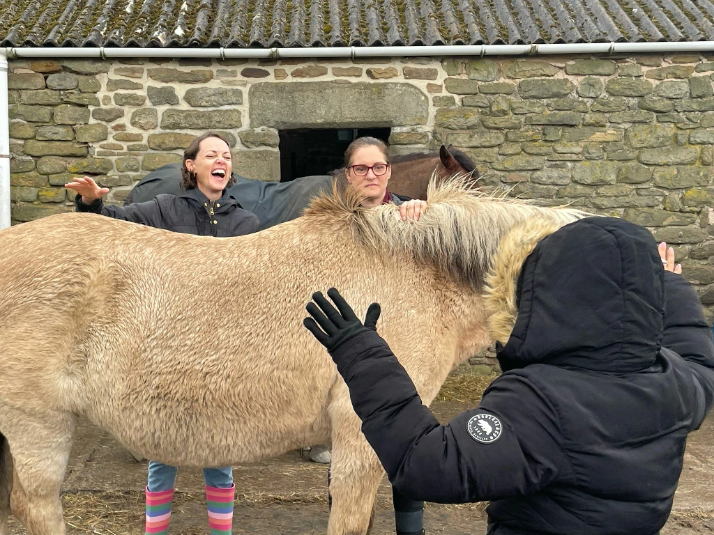 Three people petting a beige horse near a stone building.